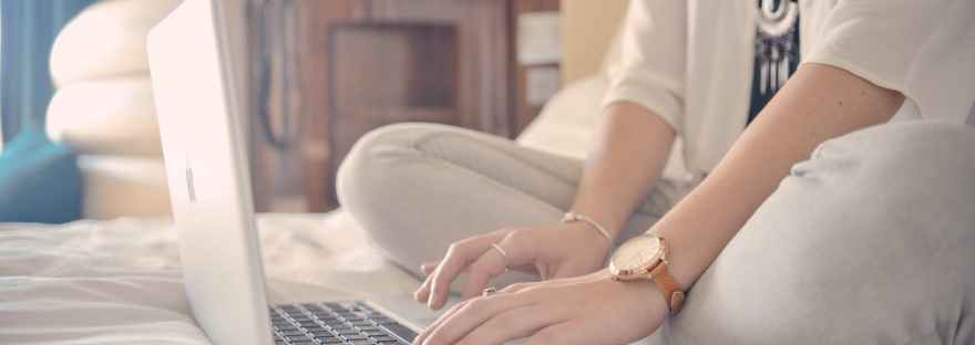 White person using a laptop on a bed, focused on hands and computer.