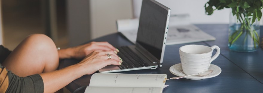 Black table with a notebook, cup of tea and laptop. A person, presumably a woman, sits casually and types.